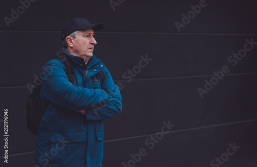 Thoughtful senior man aged 50s 60s in blue jacket and cap standing alone outdoors looking into distance, psychological mental health concept of loneliness and reflection