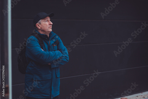 Thoughtful senior man aged 50s 60s in blue jacket and cap standing alone outdoors looking into distance, psychological mental health concept of loneliness and reflection