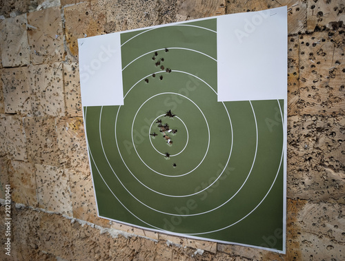 Quadro su tela Used green paper shooting target with multiple bullet holes from various pistol and rifle calibers, pinned to a wooden backstop at an indoor firing range