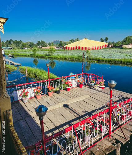 Foto The wooden sundeck of a houseboat on Dal Lake, lined with potted red geraniums, a large cafe umbrella, and a view over lily-pad-covered water toward distant hills