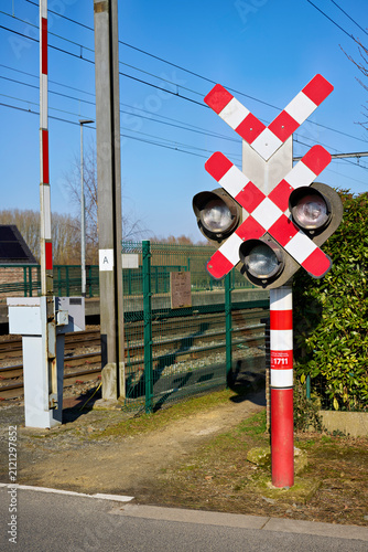 Bright outdoor shot of a railway crossing featuring a safety barrier and a distinctive St. Andrew's cross warning sign with lights, beside train tracks on a sunny day.