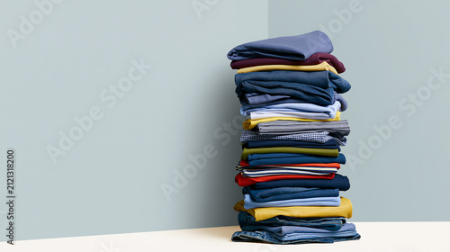 A tall stack of neatly folded colorful clothes and pants against a light blue wall corner, laundry organization and wardrobe tidy concept for clothing store display