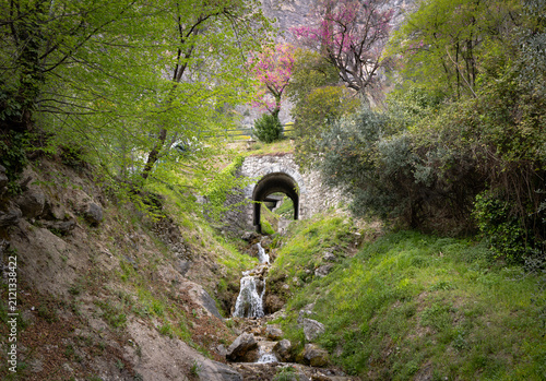 Small Mountain Waterfall Flowing Beneath a Stone Bridge in Spring, Italy