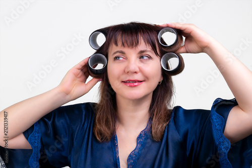 Smiling woman with curlers doing hair styling at home in blue robe