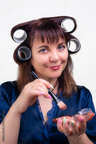A woman in curlers applies makeup with a brush, morning home ritual