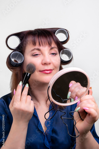 A woman with curlers applies blush in front of a mirror at home