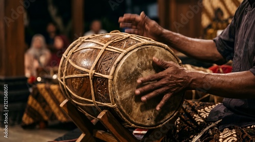 Close up of a musician's hands playing a traditional carved wooden kendang drum during an Indonesian cultural performance, wearing batik.