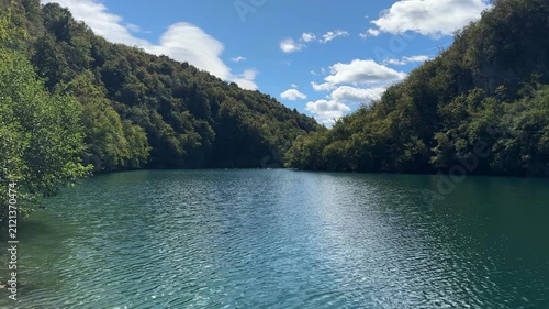 Rastovaca, Croatia - October 06, 2025: Plitvice Lakes National Park. Serene lake surrounded by lush green hills under a bright blue sky with fluffy clouds reflecting on water
