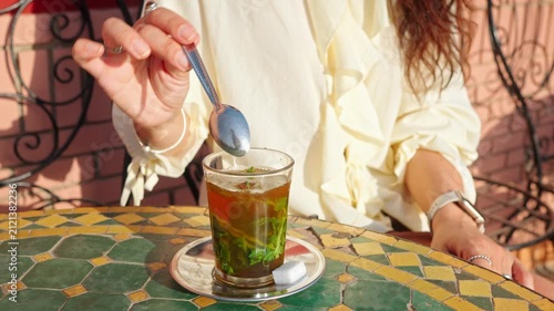 Woman Stirring Moroccan Mint Tea in Glass at Mosaic Table in Sunny Outdoor Cafe