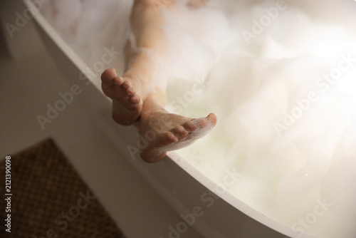 Legs and feet are resting over rim of white freestanding bathtub with dense foam, bathmat nearby