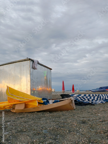 Trash on the beach.Folded beach umbrella and discarded equipment near metal booth on pebble beach with copy space cloudy sky.