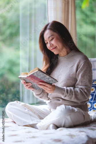 Vertical portrait image of a woman reading a book on the bed at home