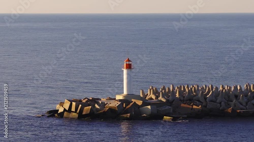 Aerial view shows a white lighthouse with red lantern on a concrete pier by tetrapod defenses off Ventspils, Latvia. Gentle ripples move under warm low light.