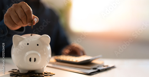 Close-up Hand putting coins into a white piggy bank saving bank for account save money. Planning step up, saving money for future plan, retirement fund. Business investment-finance accounting concept.