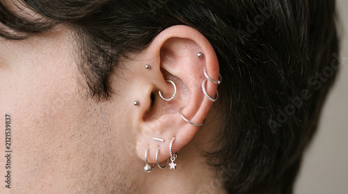 Close-up portrait of a man's ear featuring multiple silver hoop earrings and cartilage piercings, showcasing modern layered ear jewelry styling. Soft lighting highlights helix, tragus, and lobe pierci