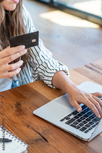 Woman holding a credit card while typing on a laptop, making an online purchase and performing e-commerce transactions on a wooden table