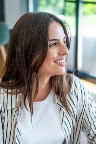 Young beautiful woman with long brown hair smiling and looking away, expressing happiness and confidence while relaxing indoors in a casual setting
