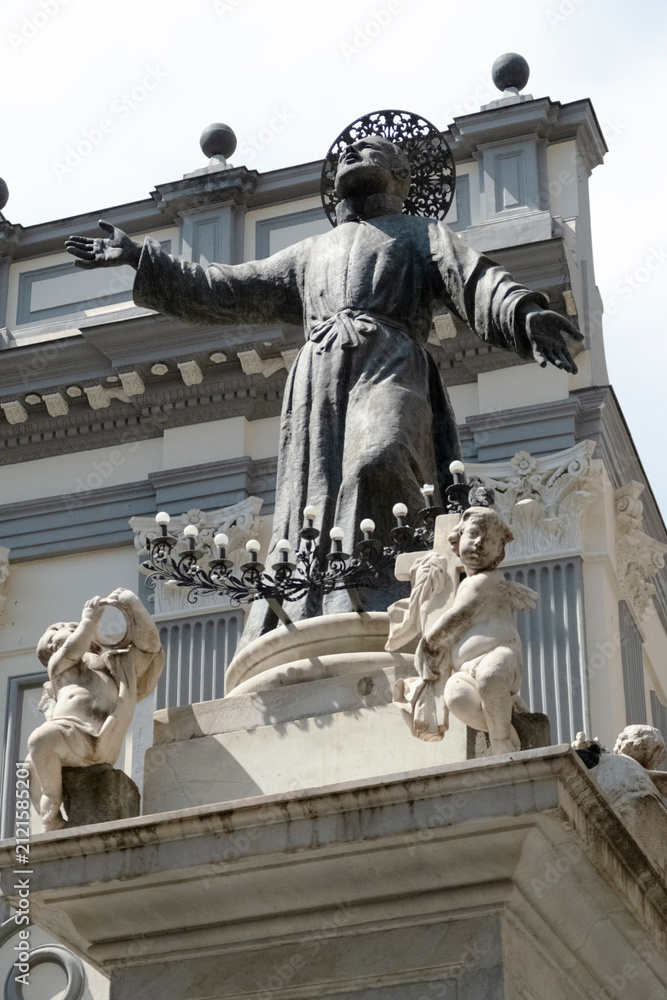 Obraz premium Ornate stone statue of Saint Francis of Assisi with halo, arms outstretched, on a church facade in Naples, Italy. Baroque architectural details frame the religious sculpture.