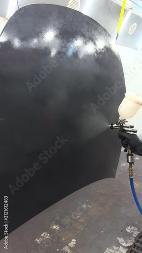 Professional auto body painter applying black primer with spray gun in booth