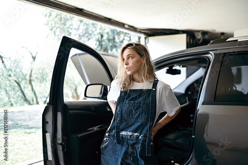 Woman in overalls stands near car in garage during daytime