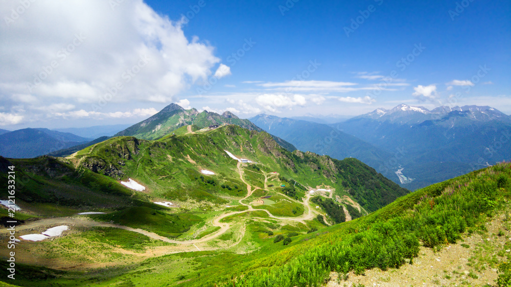 Fototapeta premium Wide landscape view of a green mountain range in summer, Rosa Khutor resort, Sochi, Russia. Winding dirt road and trails on alpine hills with patches of snow. Blue sky over distant peaks