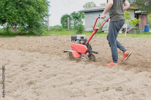 A young Caucasian man with short hair operates a red tiller in a garden. The ground is freshly tilled, surrounded by greenery and a shed in the background.
