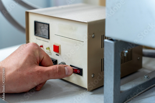 Close-up of a human hand pressing a red rocker power switch on a electrical control unit in a workshop or laboratory setting.