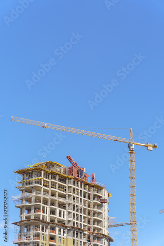 A tall yellow tower crane operating at a high-rise residential building construction site. A multi-story concrete framework of the developing skyscraper with scaffolding and insulation layers.