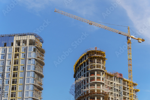 A tall yellow tower crane operating at a high-rise residential building construction site. A multi-story concrete framework of the developing skyscraper with scaffolding and insulation layers.