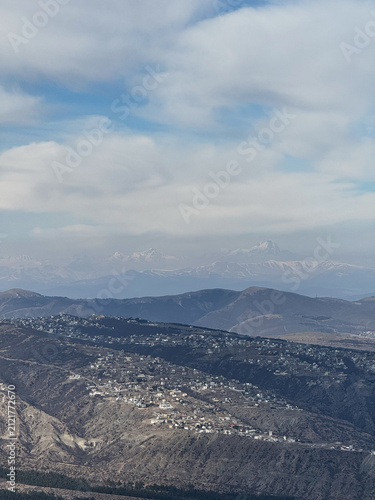 Mountain ridges with distant snow peaks and settlements under cloudy blue sky.