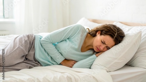 A young Caucasian woman with brown hair lies in bed, holding her stomach in discomfort. She appears to be experiencing pain or illness.