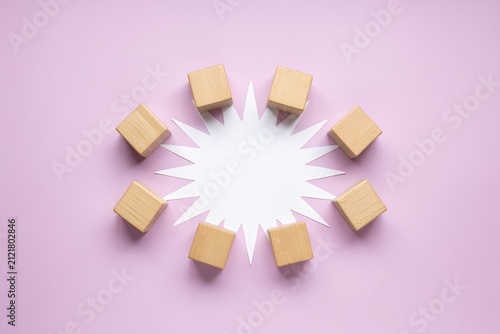 Circular arrangement of eight wooden cubes surrounding a white starburst paper cutout on a light purple background flat lay concept
