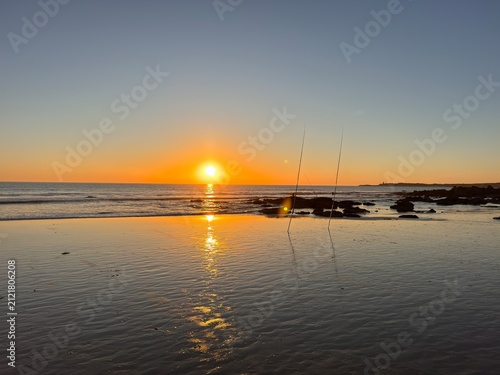 Beautiful sunset on the beach with a few rock formations and two fishing rods standing upright in the foreground, Playa de La Fontanilla in Conil de la Frontera, Spain