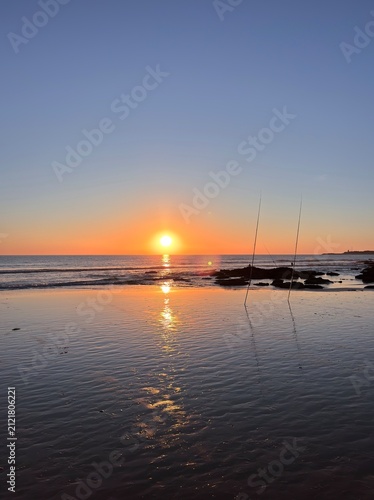 Beautiful sunset on the beach with a few rock formations and two fishing rods standing upright in the foreground, Playa de La Fontanilla in Conil de la Frontera, Spain