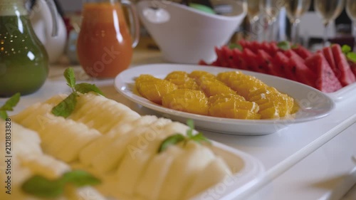 Vibrant fruit platter featuring watermelon, orange, melon slices arranged on white plates with mint leaves. Soft lighting highlights juicy textures bright colors