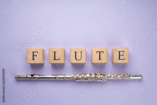 Silver flute instrument placed next to wooden letter blocks spelling flute on a light purple background