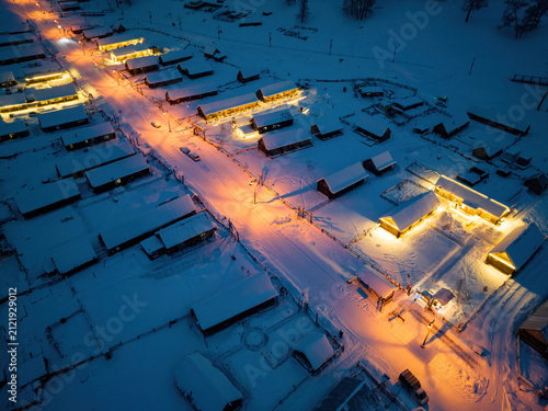 Bild auf Leinwand Aerial view of a snow-covered rural village at night, with lit buildings lining a snow-laden street, set in winter