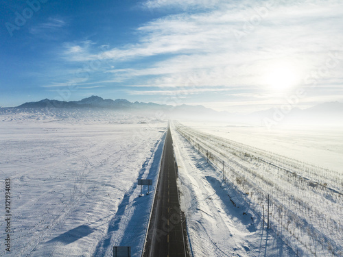 A long straight road cuts through a vast snowy plain under a clear blue sky, with distant mountains on the horizon.