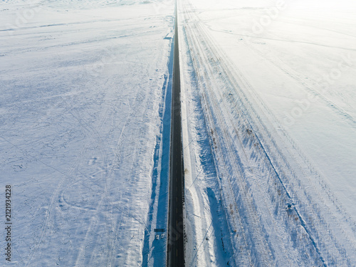 A straight highway cuts through vast snow-covered rural fields in cold winter, extending to the distant horizon.