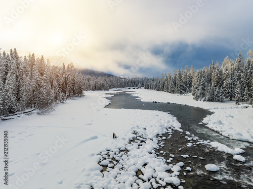 Bild auf Leinwand A winding river flows through a snow-covered coniferous forest in winter, with a small figure on the snowbank under soft sunlight