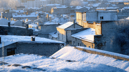 Snow-covered rooftops and buildings in a city at dawn, with the sun casting a warm glow on the snow.