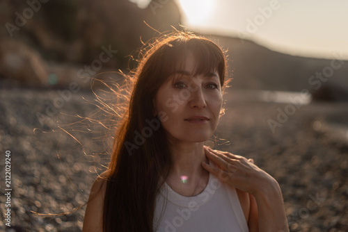 Woman, sunset, beach. Serene middle-aged woman enjoying golden hour light on a tranquil pebble shore, her hair illuminated.