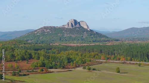 AERIAL: Scenic rocky mountain rising above forests, green fields and colourful autumn vegetation near Otočac, Croatia. Clear blue sky and layered hills create a broad, picturesque rural landscape.