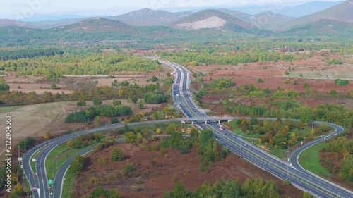 AERIAL: Large highway interchange surrounded by fields and colorful autumn woodland in Croatia. Curving ramps, bridges and moving vehicles cross a broad rural valley beneath layered mountain ridges.