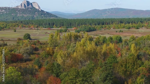 AERIAL: Rocky peak overlooking an autumn valley near Otočac, Croatia. Colorful woodland, open pasture, farmland and distant hills form a layered rural panorama beneath a pale blue sky.