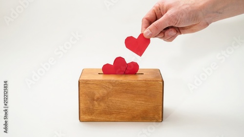 Hand places paper heart into wooden donation box with other hearts, representing charity, generosity, and acts of kindness.