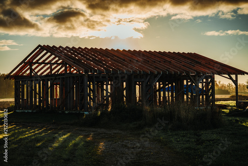 Timber-framed house under construction silhouetted against a dramatic sunset sky with exposed wooden roof trusses at a rural building site.