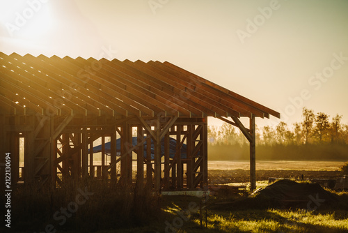 Warm golden sunlight illuminates the timber frame and roof trusses of a house under construction on a rural residential building site.