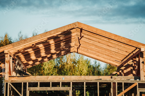 Timber frame house under construction with a Christmas wreath on the roof ridge, illuminated by warm evening sunlight in a rural countryside setting