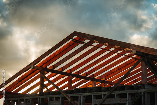 Wooden roof trusses of a timber-framed house under construction photographed against dramatic clouds during golden hour.